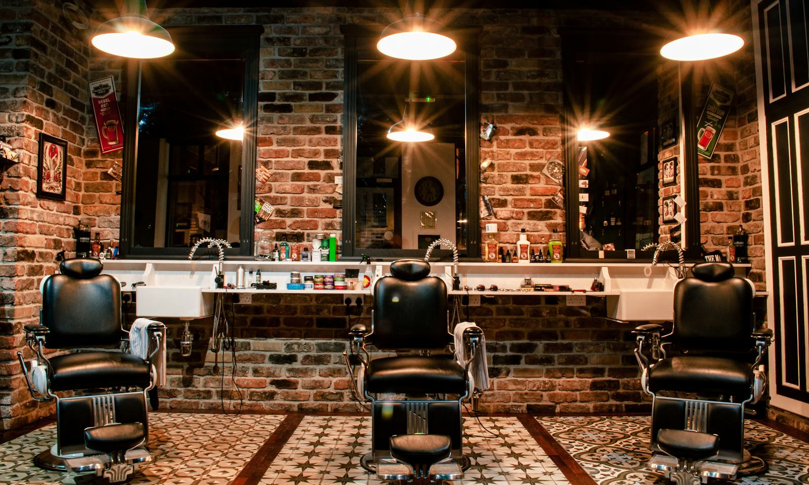 Three leather barber chairs in front of lit mirrors and a brick wall